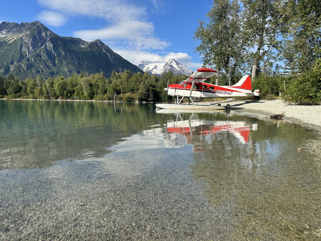Red and white floatplane parked on the gravel beach of a clear mountain lake